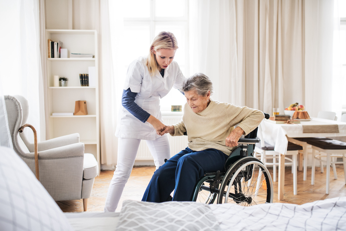 24-stunden-pflege_frau_steht_aus_rollstuhl_auf Pflegerin unterstützt Seniorin aus dem Rollstuhl aufzustehen.